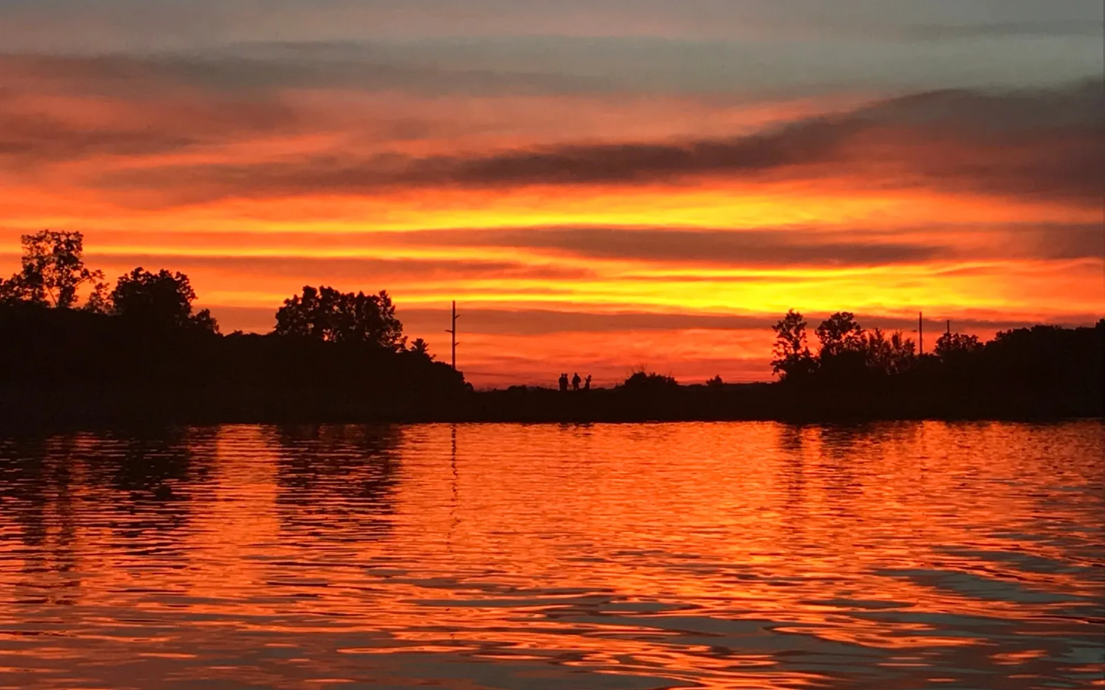 A fiery orange sunset over Lake Bella Vista, with trees silhouetted along the shore and the glowing sky rippling across the water.