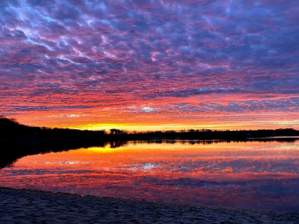 A pink, violet and orange sunset sky reflected in still Silver Lake, seen from a sandy shoreline.